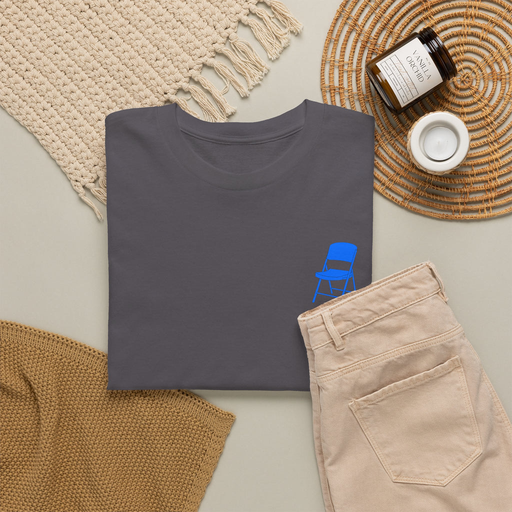 Dark gray t-shirt with blue chair graphic on a light surface with beige pants and decorative items.